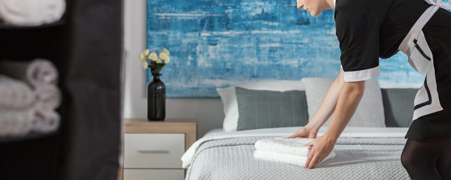 Housekeeper arranging folded white towels on a hotel bed in a modern room with blue artwork.