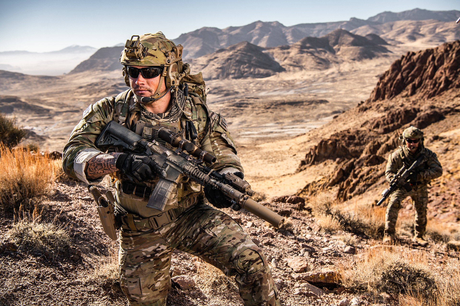 Armed soldier in camouflage uniform crouches in a desert terrain while aiming a rifle with scope, with another soldier visible in the background.