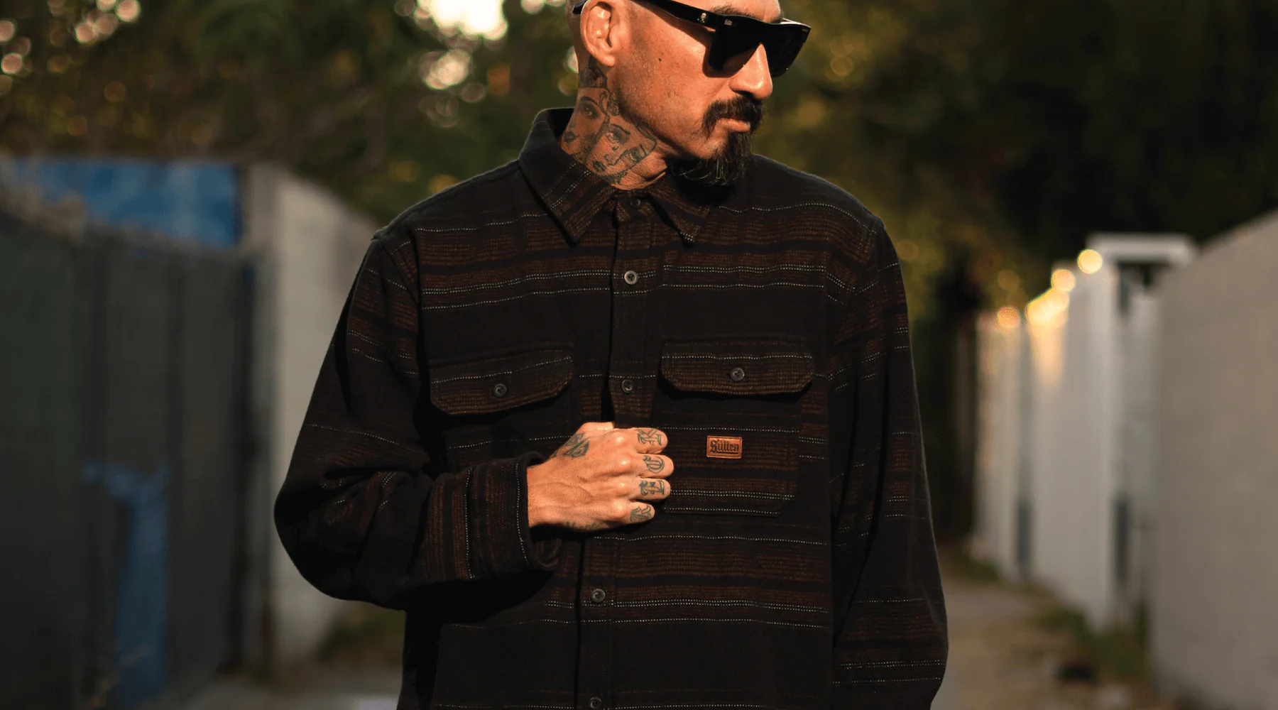 Man with tattoos and sunglasses wearing a dark striped shirt with an orange label standing outdoors in an evening setting.