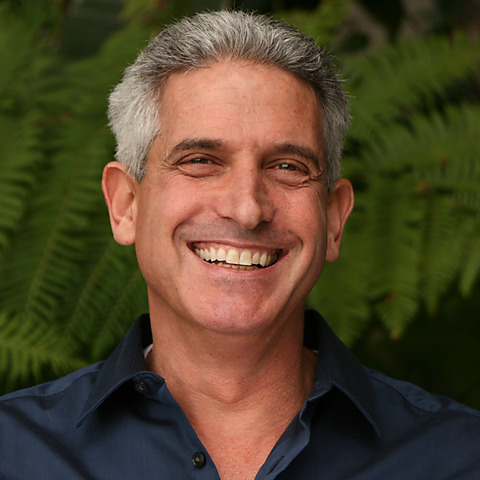 Middle-aged man with gray hair smiling at the camera while wearing a dark navy button-up shirt against a blurred green foliage background.