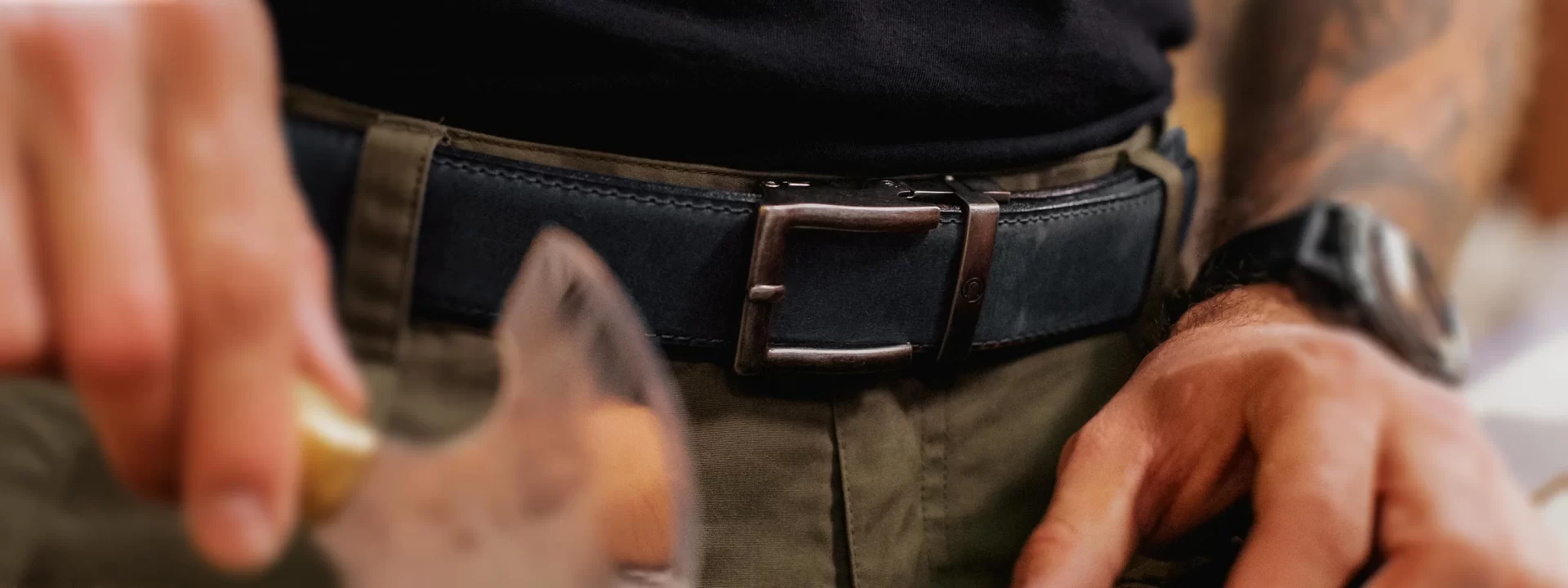 A person wearing a black belt with a bronze buckle and olive-colored pants stands with hands at their sides.