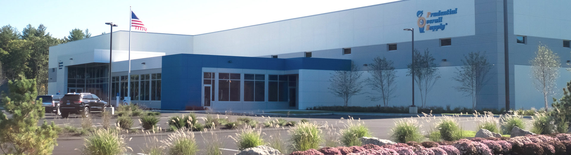 Modern commercial building with white and blue exterior, American flag, and landscaped grounds featuring ornamental grasses and shrubs.